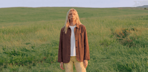 A model in a brown jacket photographed standing in a grass field