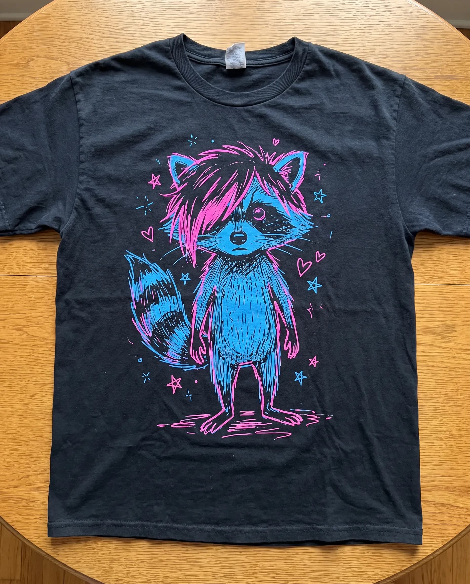 Black t-shirt flat lay with a two-color screen print of a raccoon with a pink-streaked fringe, scene art style