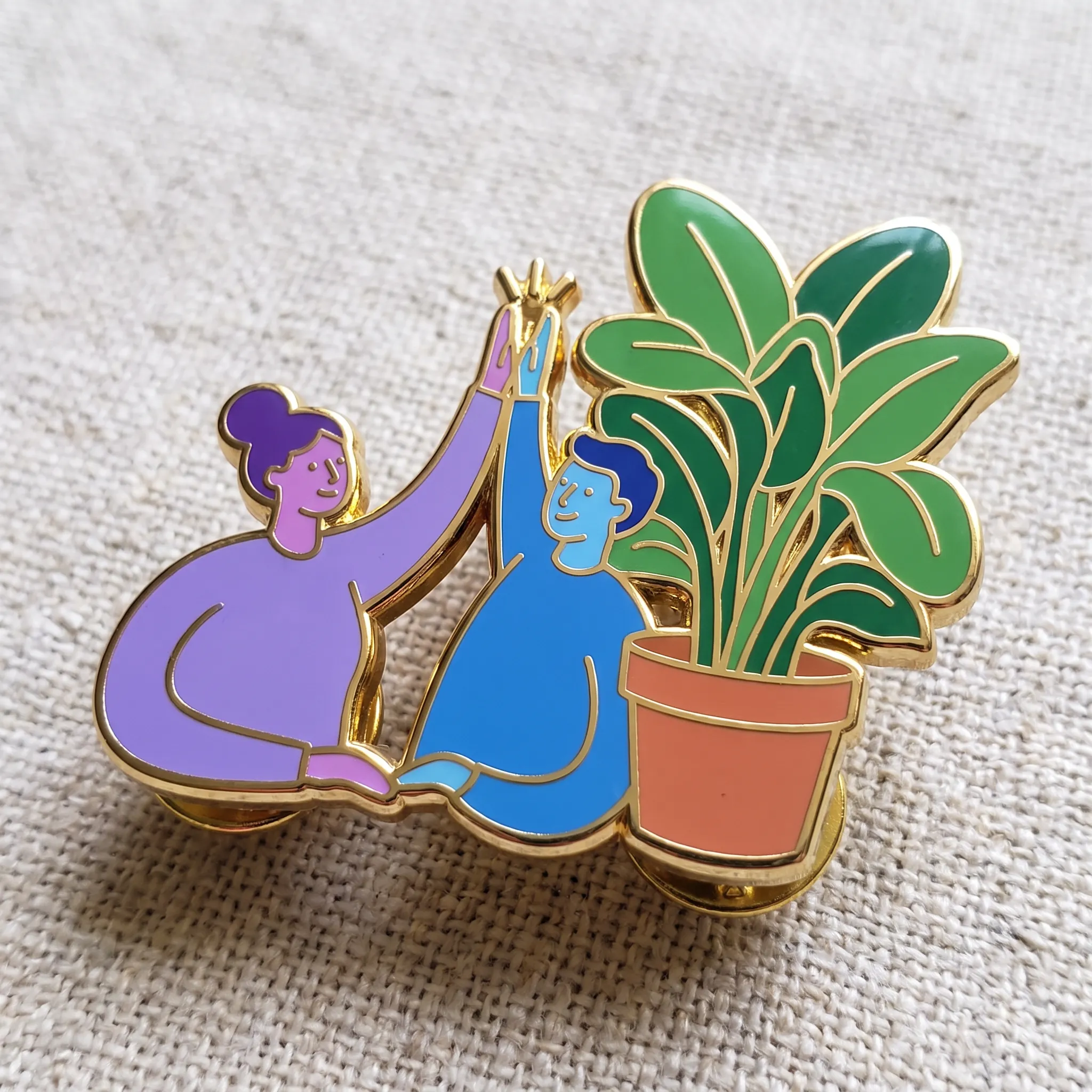 Hard enamel pin of two coworkers with purple and blue skin high-fiving beside a giant potted plant