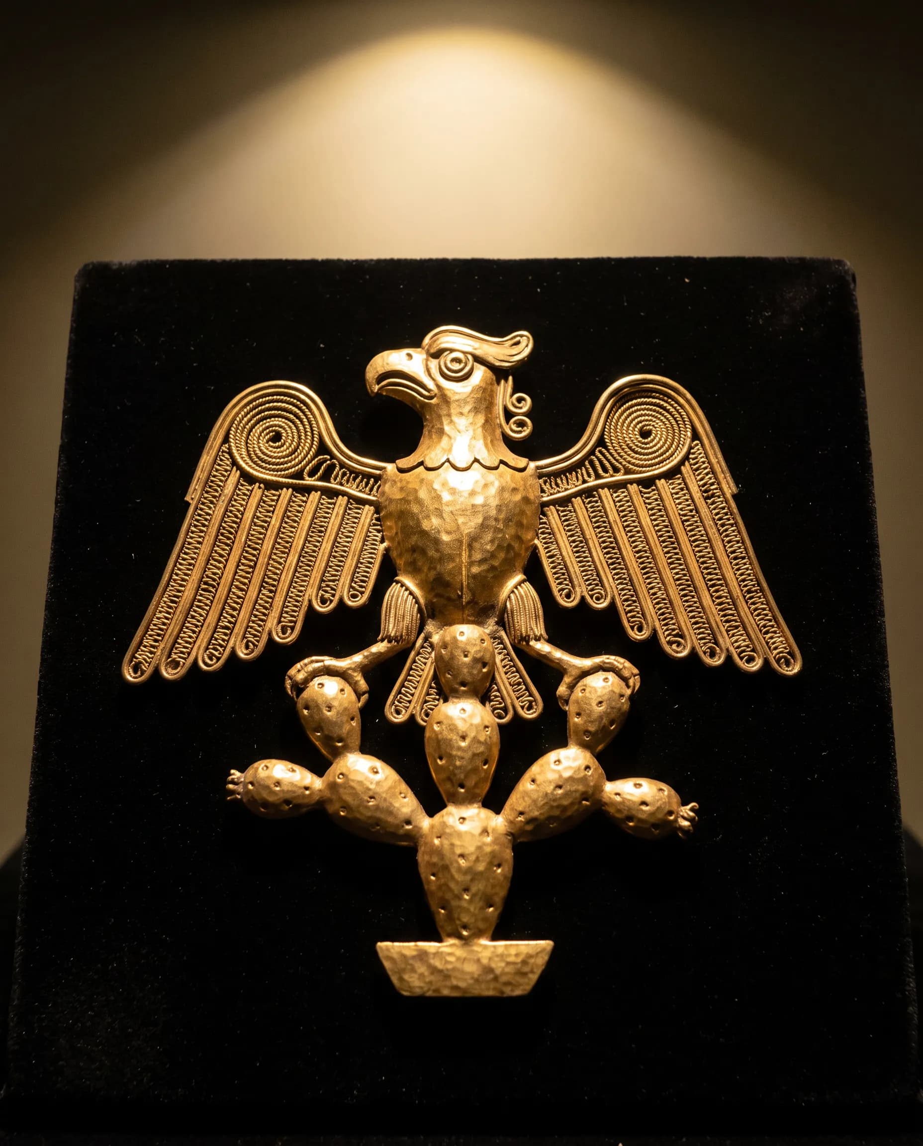 Gold ornament of an eagle perched on a nopal cactus on black velvet, Aztec art style