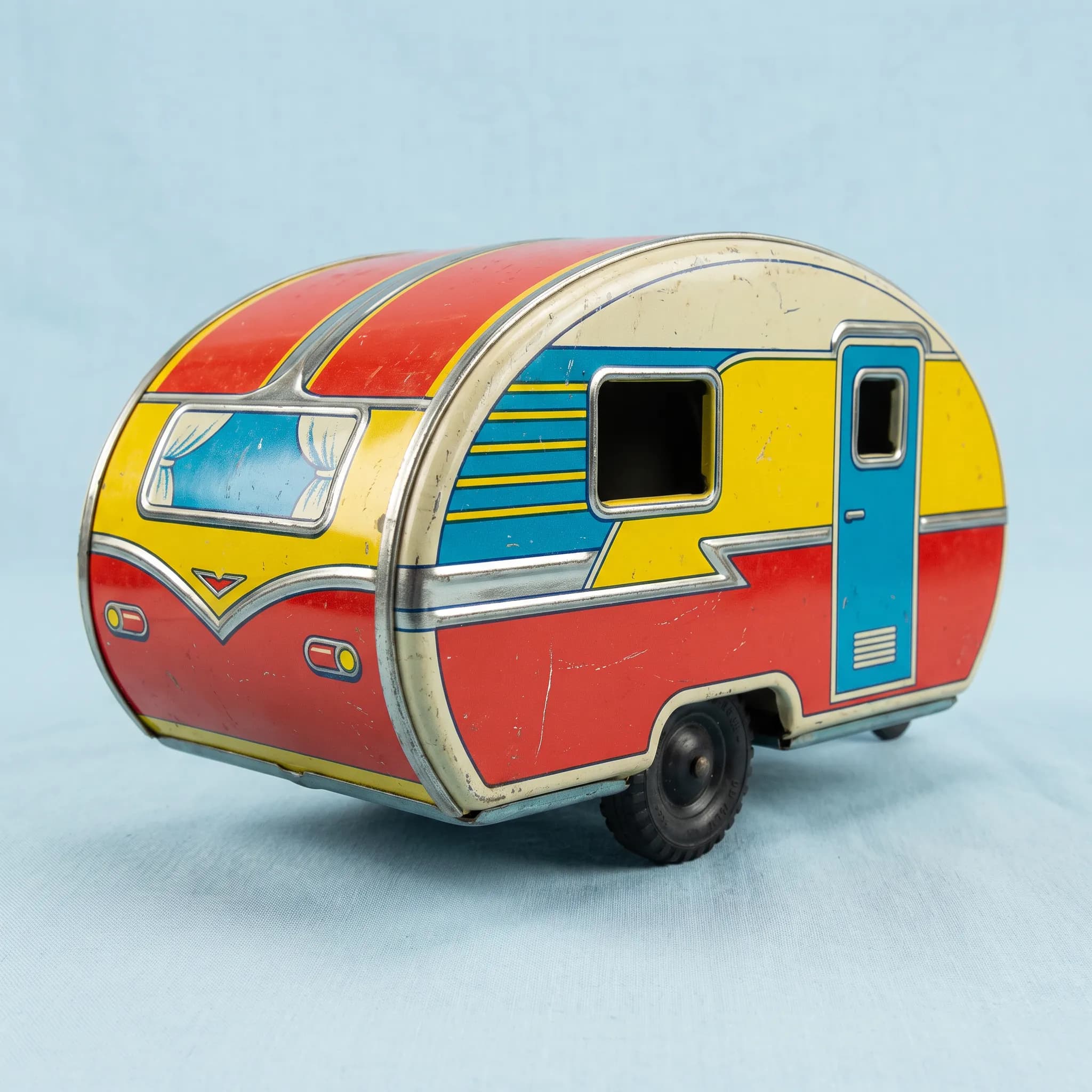 Lithographed tin toy of a teardrop camper trailer on a pale blue backdrop, retro art style